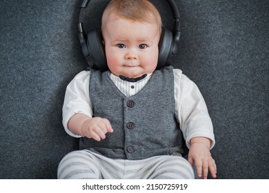 Cute Little Well Dressed Smart Baby Business Man In Vest And Bow Tie With Big Earplugs, Headphones Or Earphones Listening To Music While Holding A Phone Or Mobile For Entertainment On Grey Background