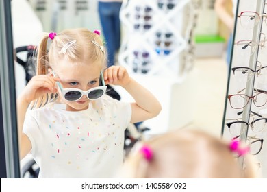 Cute Little Young Caucasian Blond Girl Trying On And Choosing Sunglasses In Front Of Mirror At Optic Eyewear Store. Adorable Schoolgirl Child Having Fun Bying Sun Uv Protection Glasses At Shop