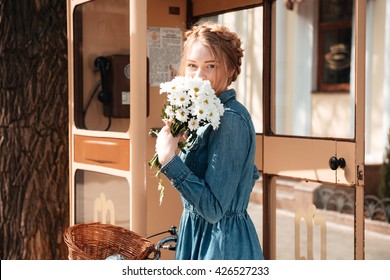 Cute lovely young woman with bicycle and bouquet of camomiles standing near telephone box on the street Stock Photo