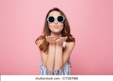 Cute lovely young woman in round sunglasses standing and sending kiss over pink background