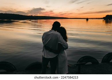 The cute loving couple standing near lake at beautiful sunset. Warm orange clouds. Silhouette evening photos. Hugging together behind back.