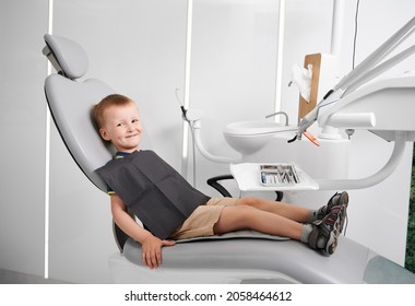 Cute Male Child Sitting In Dental Chair In Modern Clinic. Adorable Kid With Black Dental Napkin On His Chest Waiting For Procedure In Dental Office. Concept Of Pediatric Dentistry And Dental Care.