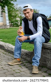 Симпатичный мужчина в повседневной одежде сидит на скамейке и ест уличную еду (шашлык)