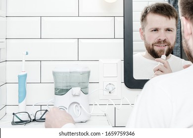Cute man cleans his teeth with the help of electric oral irrigator with a jet of water, looking in the mirror. Copy space.