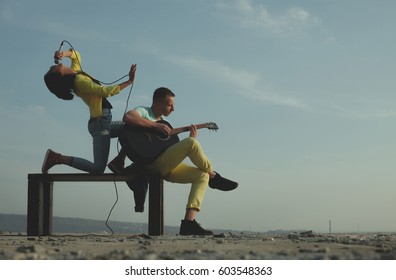 Cute Man Serenading His Girlfriend On Camping Trip On A Sunny Day. Portrait Of Young Hispanic Cute Girl And Man Plays Acoustic Guitar. Group Of Happy Friends With Guitar Having Fun Outdoor. 