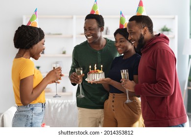 Cute Millennial Black Friends Making Surprise For Birthday Woman, Holding Cake With Lit Candles, Wearing Party Hats And Smiling, Greeting Emotional African American Lady, Side View, Home Interior