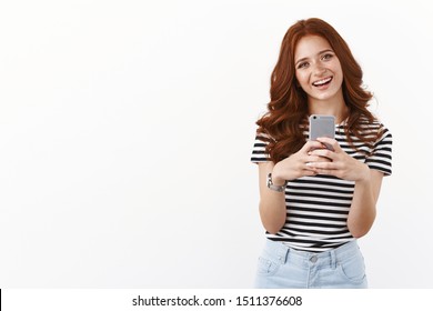 Cute modern millennial girl with red curly hairstyle, tilt head joyfully, smiling enthusiastic, holding smartphone, taking selfie in mirror, making goofy grin, messaging friends, white background
