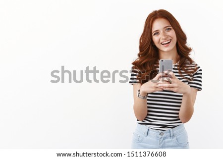 Cute modern millennial girl with red curly hairstyle, tilt head joyfully, smiling enthusiastic, holding smartphone, taking selfie in mirror, making goofy grin, messaging friends, white background