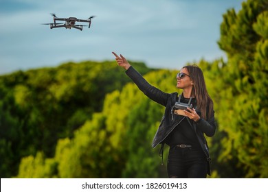 Cute Modern Smiling Woman Pilot In Dark Stylish Clothes Flying Drone In Park At Sunset. Outdoors Hobby.