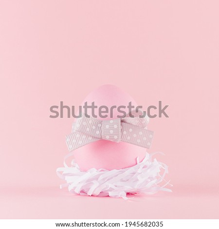 Cute naivety easter background with pink egg with grey bow in white nest on pastel pink backdrop, square.