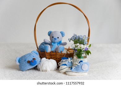 Cute Newborn Baby Boy Fashion Set Of Blue Booties, Teddy Beanie Hat And Stuffed Teddy Bear Toy Handmade With Crochet Technique From Wool On A Fluffy Blanket With Flower Bouquet And Wooden Basket