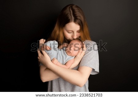 A cute newborn baby boy sleeps in a blue overalls in the first days of life. In the arms of the mother. Warm motherly hugs. On a black background. Professional portrait photography.