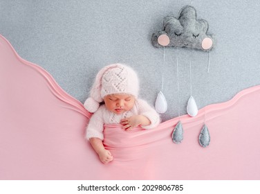 Cute newborn baby girl in knitted hat sleeping on grey stylized background with rainy cloud. Adorable infant child napping under pink blanket during studio photoshoot