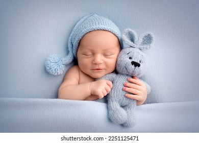A cute newborn boy in the first days of life sleeps naked on a blue fabric background. The kid gently hugs a light blue knitted bunny. Studio professional macro photography, newborn baby portrait.