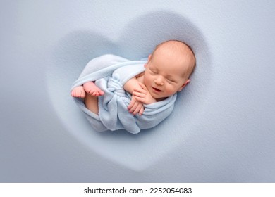 A cute newborn boy in the first days of life sleeps naked on a blue fabric background. The baby is wrapped in a blue winding, a blanket. The child lies in a mold in the shape of a heart. 