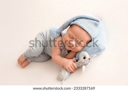 A cute newborn boy in the first days of life sleeps in a blue overalls against a background of white fabric. Blue knitted hat with pouffe. Blue head pillow. With felt gray toy giraffe.