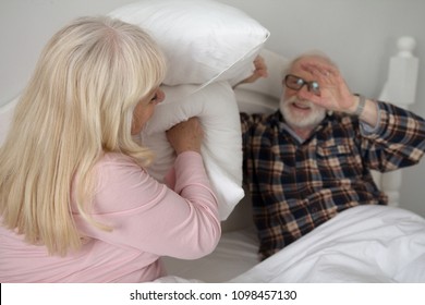 Cute Old Man And Woman Pillow Fighting In Their Bed. Sweet Senior Couple Wearing Pajamas Being Silly Having Pillow Fight In Bedroom.