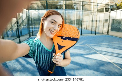 A cute paddle tennis player takes a picture of herself happily posing with a paddle tennis racket. Outdoor paddle tennis concept, women playing paddle tennis.