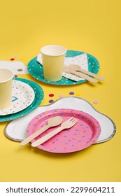 Cute paper party plates and cups for themed kids party on yellow background. Birthday party decorations and props in bright colors. Set of holiday disposable tableware for party or picnic. 