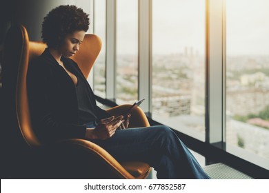 Cute pensive afro american student is sitting on armchair in front of window of skyscraper and having online conversation with her curator during probation period in office via digital tablet