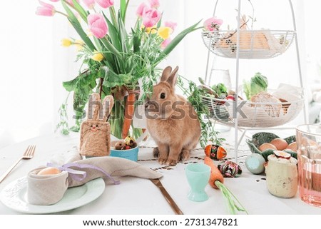 Cute pet rabbit on Easter table with fresh flowers, carrots and painted Easter eggs