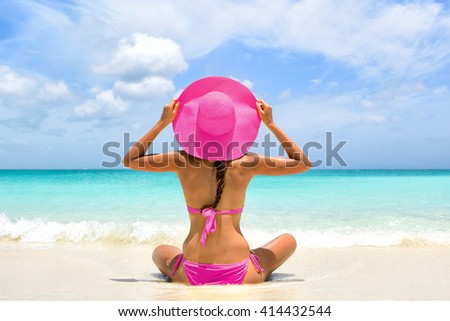 Cute pink bikini beachwear woman relaxing in perfect paradise destination on beach travel vacation. Girl from the back holding fashion straw floppy hat sitting on sand looking a turquoise ocean.