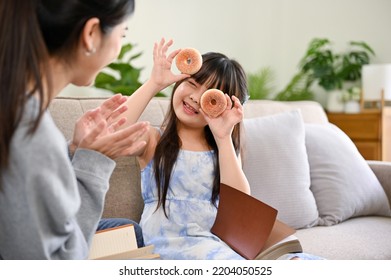 A Cute And Playful Young Asian Girl Holding Doughnuts, Playing Peekaboo With Her Mom While Relaxing In The Living Room Together. Happy Family Moments Concept.