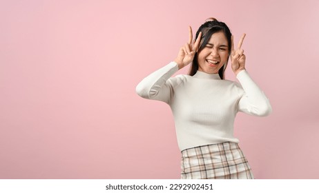 Cute and playful young Asian woman in casual clothes showing two fingers, posing peace sign, isolated on pink background. - Powered by Shutterstock