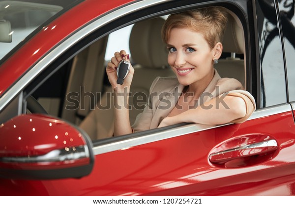 Cute pleased blonde business woman in a red car\
holding car key in her hand and looking at camera. Car Rent or Auto\
purchase Concept.