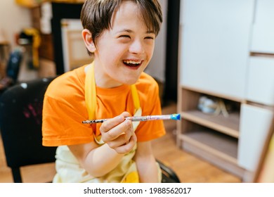 Cute positive glückliche Junge mit Down-Syndrom sitzend in einem Stuhl und Zeichnung mit Pinsel drinnen. Konzeption des Lernens behinderter Kinder.