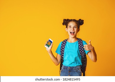 Cute positive schoolgirl 10 years old in casual clothes with backpack, sitting on yellow background, in Studio, holding mobile phone. Dependence on gadgets and Internet. E-education. Copy space.