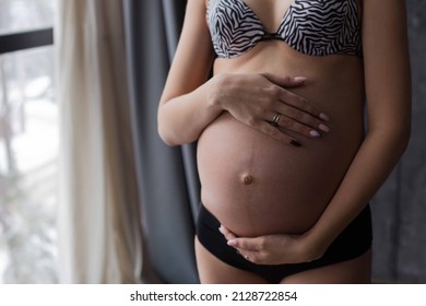 Cute pregnant belly isolated on the background. A view of a young pregnant woman hugging her bare stomach with her hands. Big belly in the third trimester of pregnancy close-up. The concept of pregnan