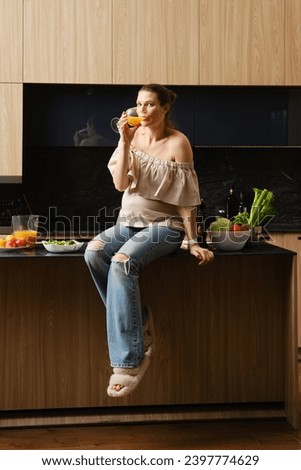 Cute pregnant woman drinks juice sitting on kitchen tabletop