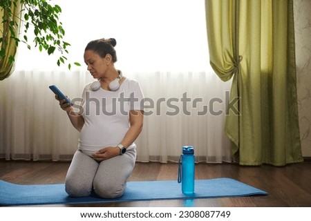 Cute pregnant woman sitting in hero pose, gently stroking her belly, adjusting soundtrack on mobile app on her smartphone, training at home, practicing stretching exercises and pregnancy yoga. 9 month