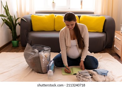 Cute Pregnant Woman Writing Packing List For Maternity Hospital Notebook Prepares Bag. Young Ledy In Pregnancy Have Fun Spend Time On Floor Near Sofa At Home. Motherhood, Medicine Health Care Concept
