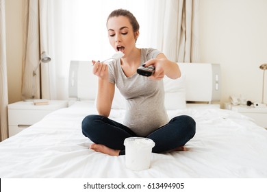 Cute pregnant young woman with remote control eating ice cream and watching TV at home
