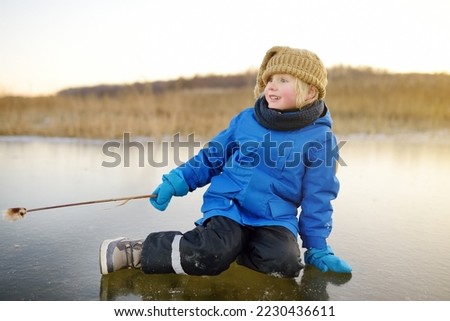 Cute preschool boy is playing on the ice of a frozen lake or river on a cold sunny winter sunset. Child having fun with icicle and dry reed plant during family hiking. Kids outdoor games in winter