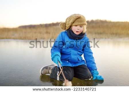 Cute preschool boy is playing on the ice of a frozen lake or river on a cold sunny winter sunset. Child having fun with icicle and dry reed plant during family hiking. Kids outdoor games in winter