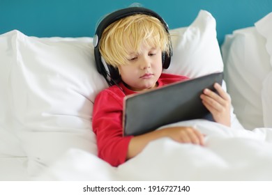 Cute Preschool Boy Wearing Headphones Watches Cartoon Movie By Tablet In Bed In The Morning Right After Waking Up. Overuse And Addiction Kids From Gadgets.