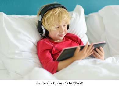 Cute Preschool Boy Wearing Headphones Watches Cartoon Movie By Tablet In Bed In The Morning Right After Waking Up. Overuse And Addiction Kids From Gadgets.