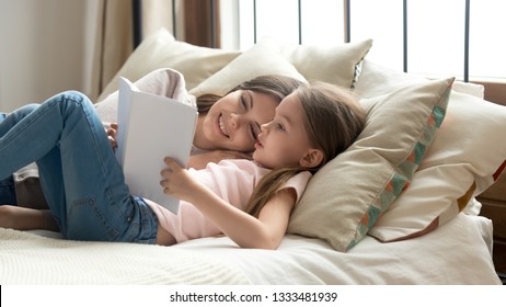 Cute Preschool Child Daughter Holding Book Reading Fairy Tale To Mom Lying In Bed Together, Happy Mother Listening Focused Little Kid Girl Learning To Read Relaxing In Bedroom Enjoy Bedtime Stories