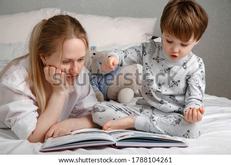 Cute preschool child son holding a book reading fairy tales for mom, lying in bed together, happy mother listening to a focused little boy learning to read, enjoying bedtime stories