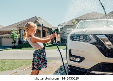 Cute Preschool Little Caucasian Girl Washing Car On Driveway In Front House On Sunny Summer Day. Kids Home Errands Duty Chores Responsibility Concept. Child Playing With Hose Spraying Water.