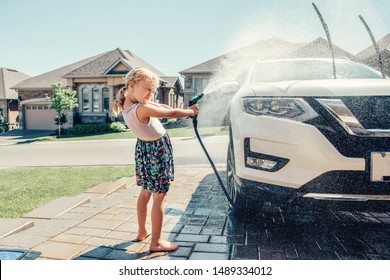 Cute Preschool Little Caucasian Girl Washing Car On Driveway In Front House On Sunny Summer Day. Kids Home Errands Duty Chores Responsibility Concept. Child Playing With Hose Spraying Water.