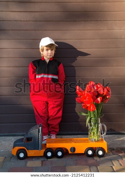 cute preschooler boy in a red overalls uniform\
and a cap stands near big toy truck with bouquet of red tulip\
flowers. Delivery concept, congratulation for mom on mother\'s day,\
birthday little courier