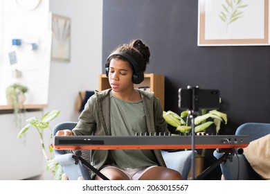 Cute preteen biracial schoolgirl in headphones playing piano keyboard while sitting in domestic room - Powered by Shutterstock