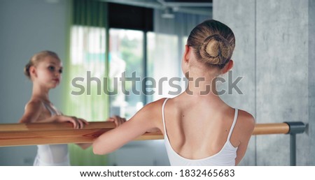 Cute preteen girl ballerina with bun in white costume looks into large mirror leaning on wooden barre in dance studio