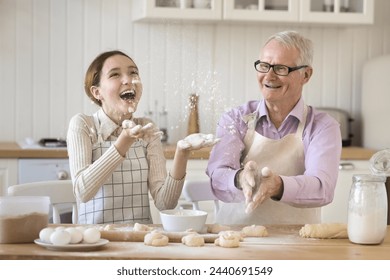 Cute preteen granddaughter in apron having fun with older grandpa sit at table cooking, throwing, sprinkling white flour while prepare homemade dessert, buns, sweets at home. Culinary, family holiday