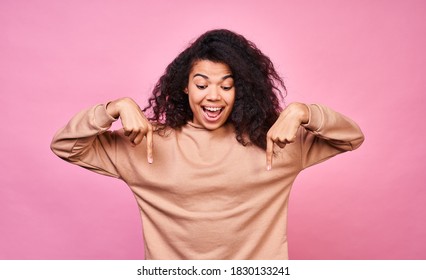 Cute pretty African American female with clean healthy skin, smiling happily, pointing her fingers at something down, isolated on pink background. A pretty young woman sees an amazing thing below. - Powered by Shutterstock