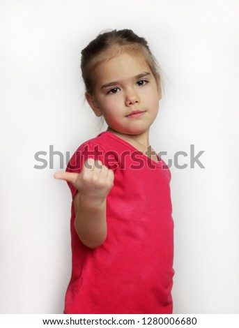 Cute pretty preschool girl in red t-shirt showing something on her right direction with thumb up gesture over white background, child emotional portrait, indoor closeup 